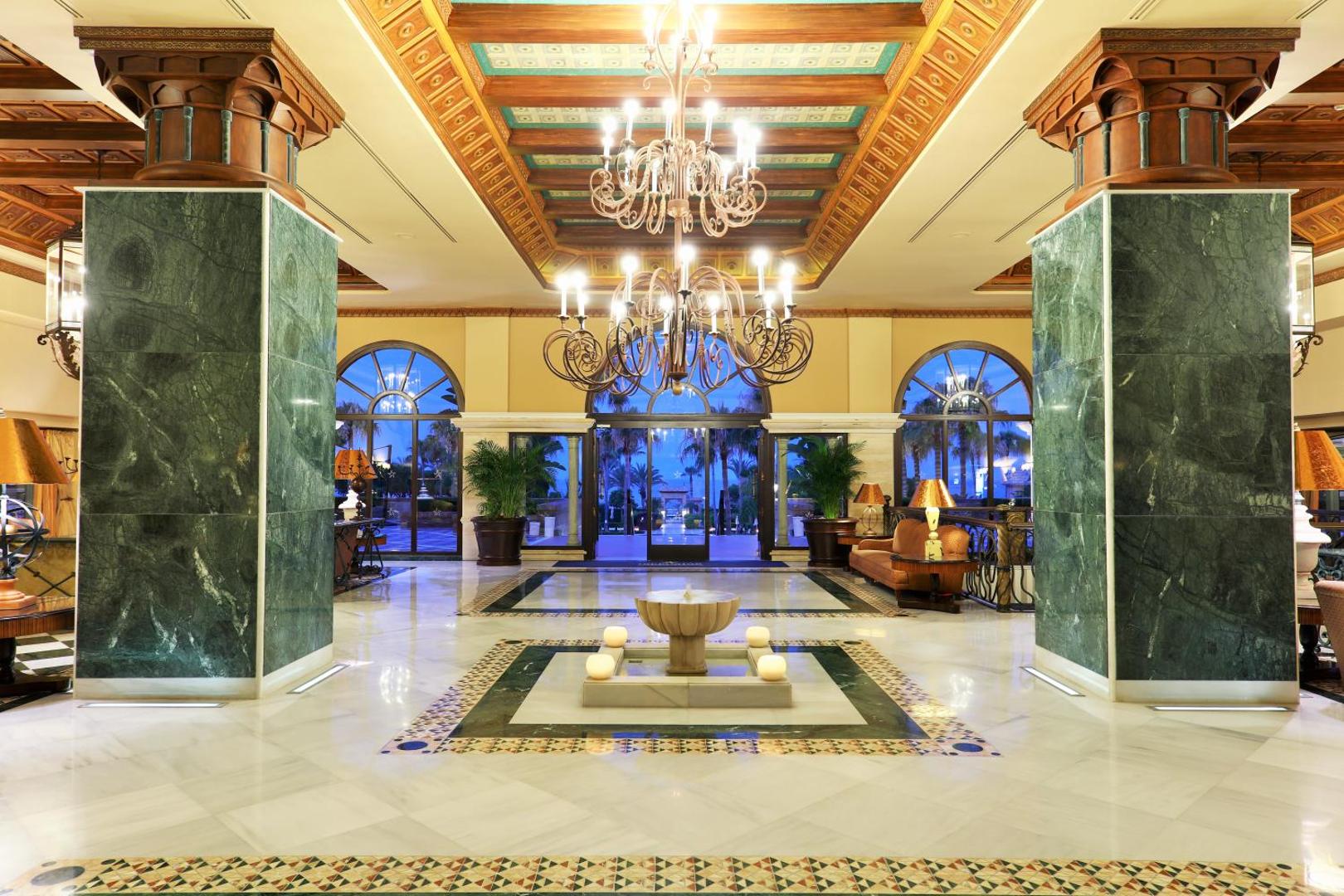 Iberostar Malaga Playa greznais vestibils ar marmora grīdām, masīvām zaļām kolonnām, lielām lustrām un skatu uz dārzu.