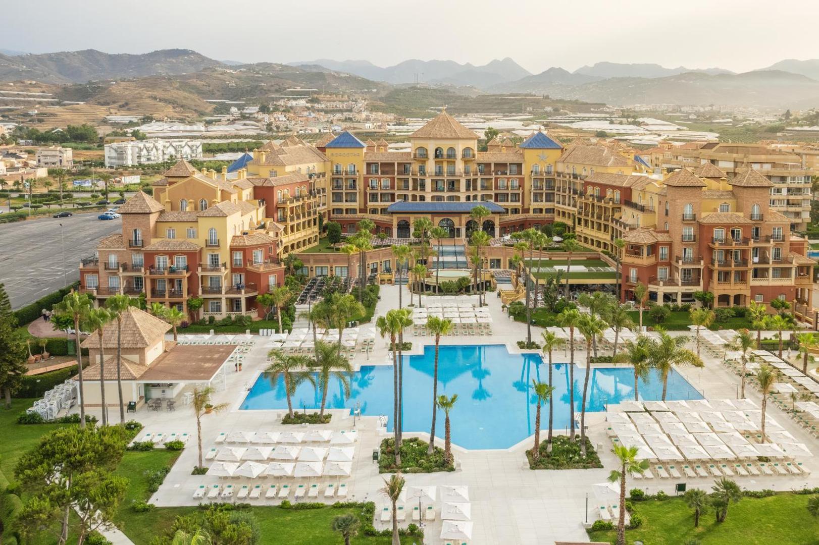 Iberostar Malaga Playa viesnīcas kopskats ar lielu āra baseinu, saulessargiem un palmu dārzu Andalūzijas kalnu fonā Spānijā