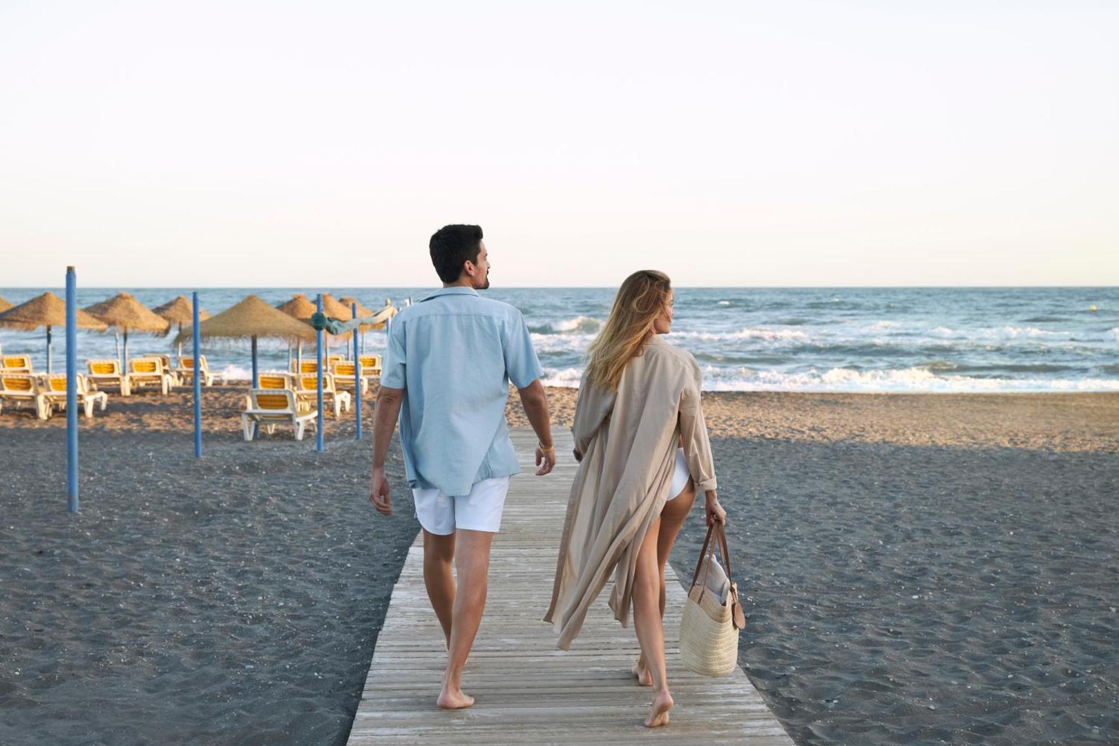 Pāris pastaigājas pa koka laipu uz Iberostar Malaga Playa privāto pludmali saulrieta laikā — fonā jūra un saulessargi