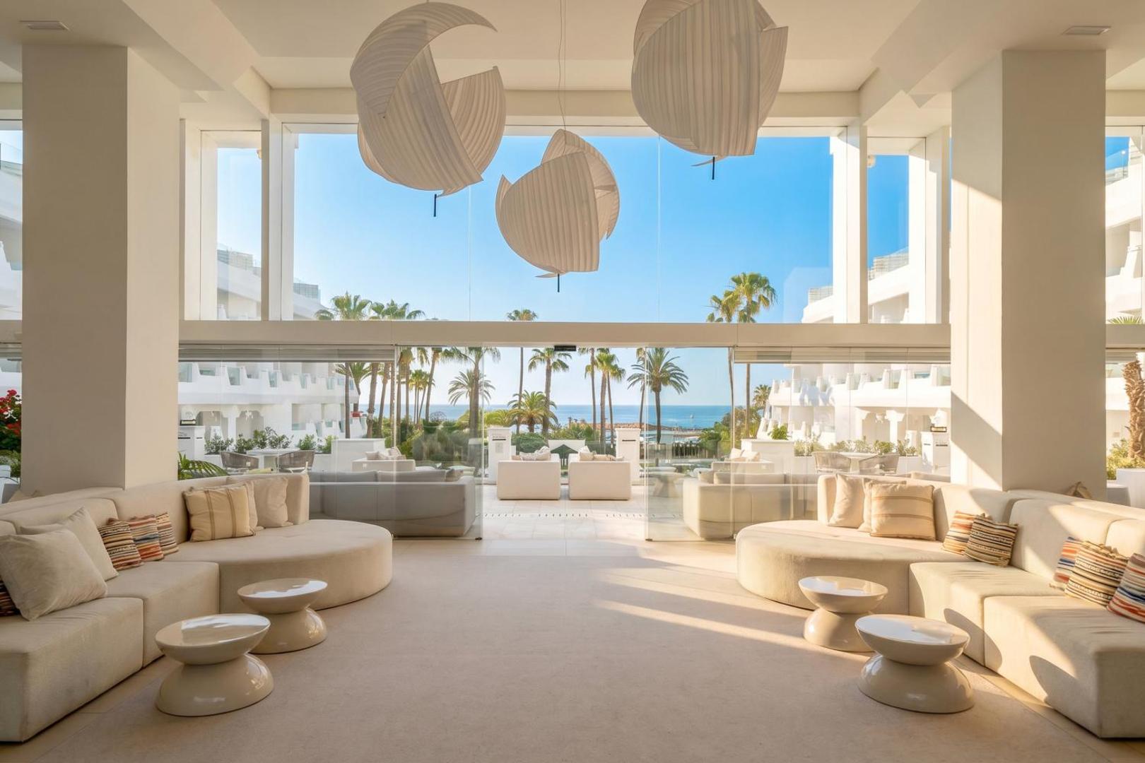 Iberostar Marbella Coral Beach viesnīcas lobby atpūtas zona ar gaišiem dīvāniem, dizaina gaismekļiem un skatu caur lielajiem logiem uz palmām un jūru