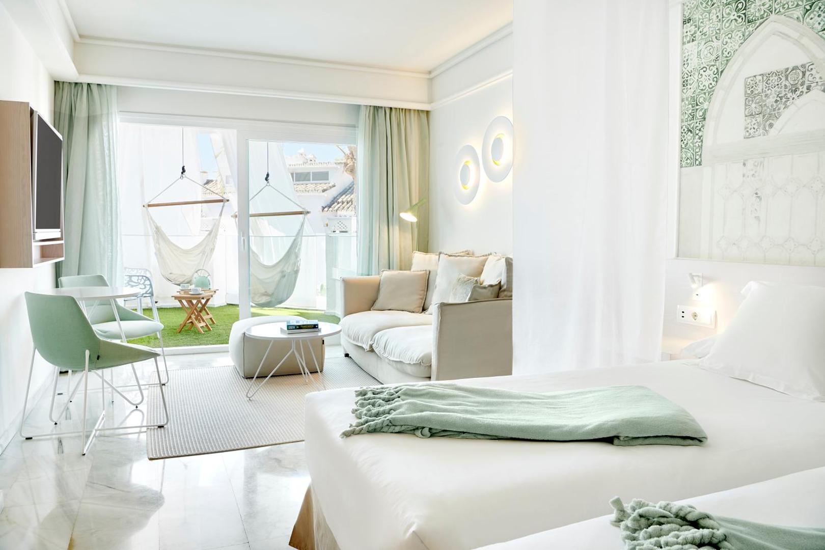 Iberostar Marbella Coral Beach gaišs viesnīcas numurs ar atpūtas zonu, dīvānu un terasi, kurā uzstādīti divi balti šūpuļtīkli virs mākslīgā zālāja.