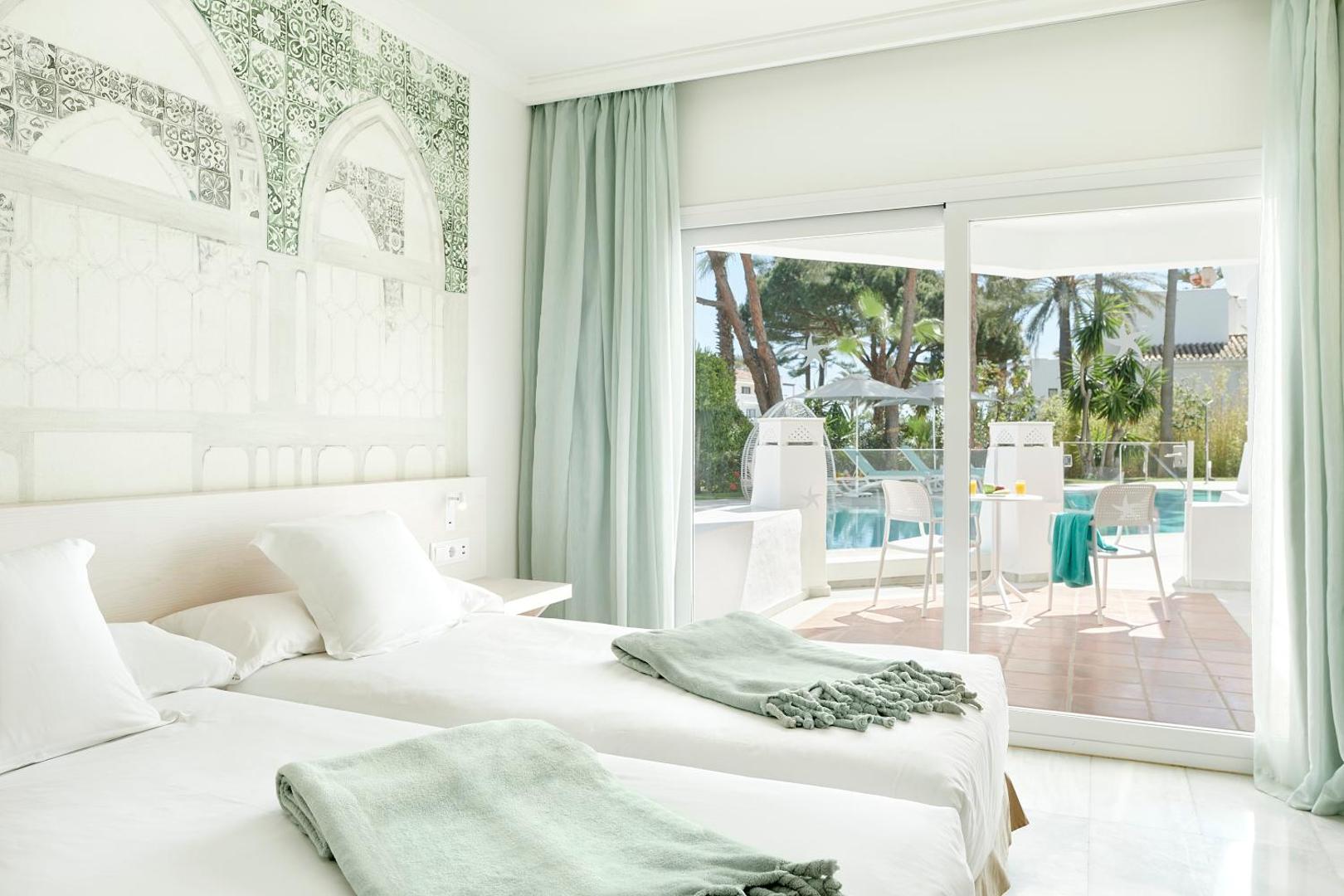 Iberostar Marbella Coral Beach numura interjers ar skatu caur stikla durvīm uz baseina zonu, āra mēbelēm un palmām saulainā dienā.