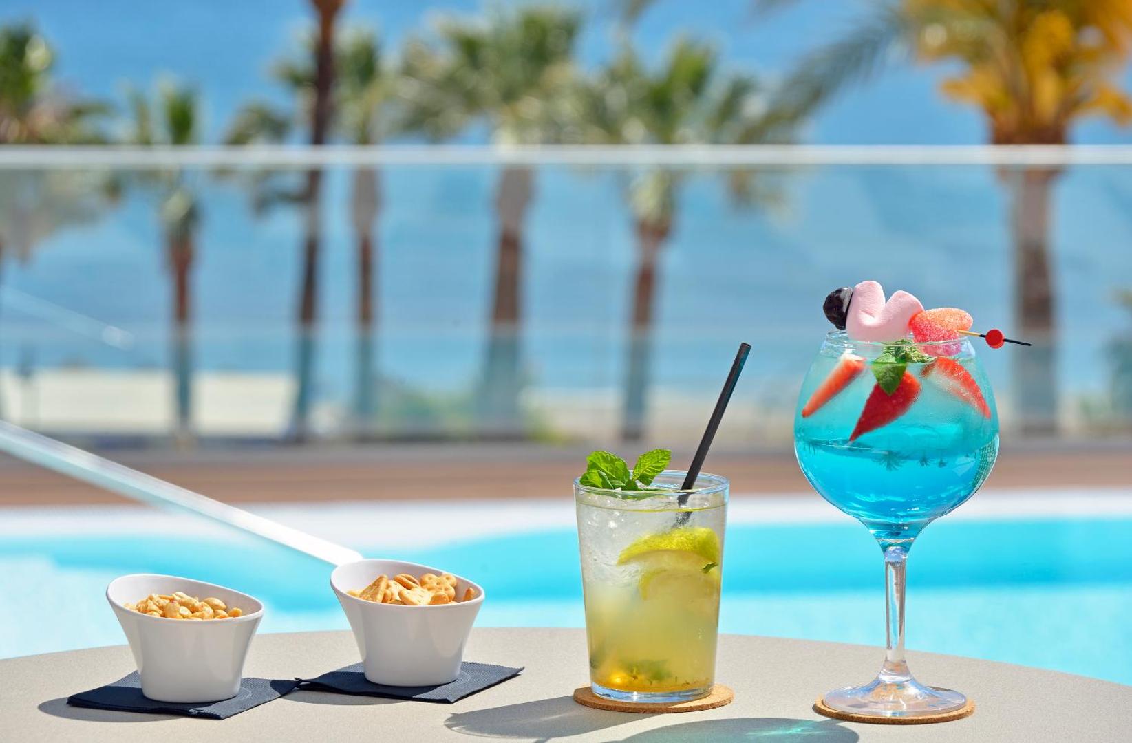 Krāsaini kokteiļi un uzkodas uz galda pie Ocean House Costa del Sol baseina ar skatu uz jūru