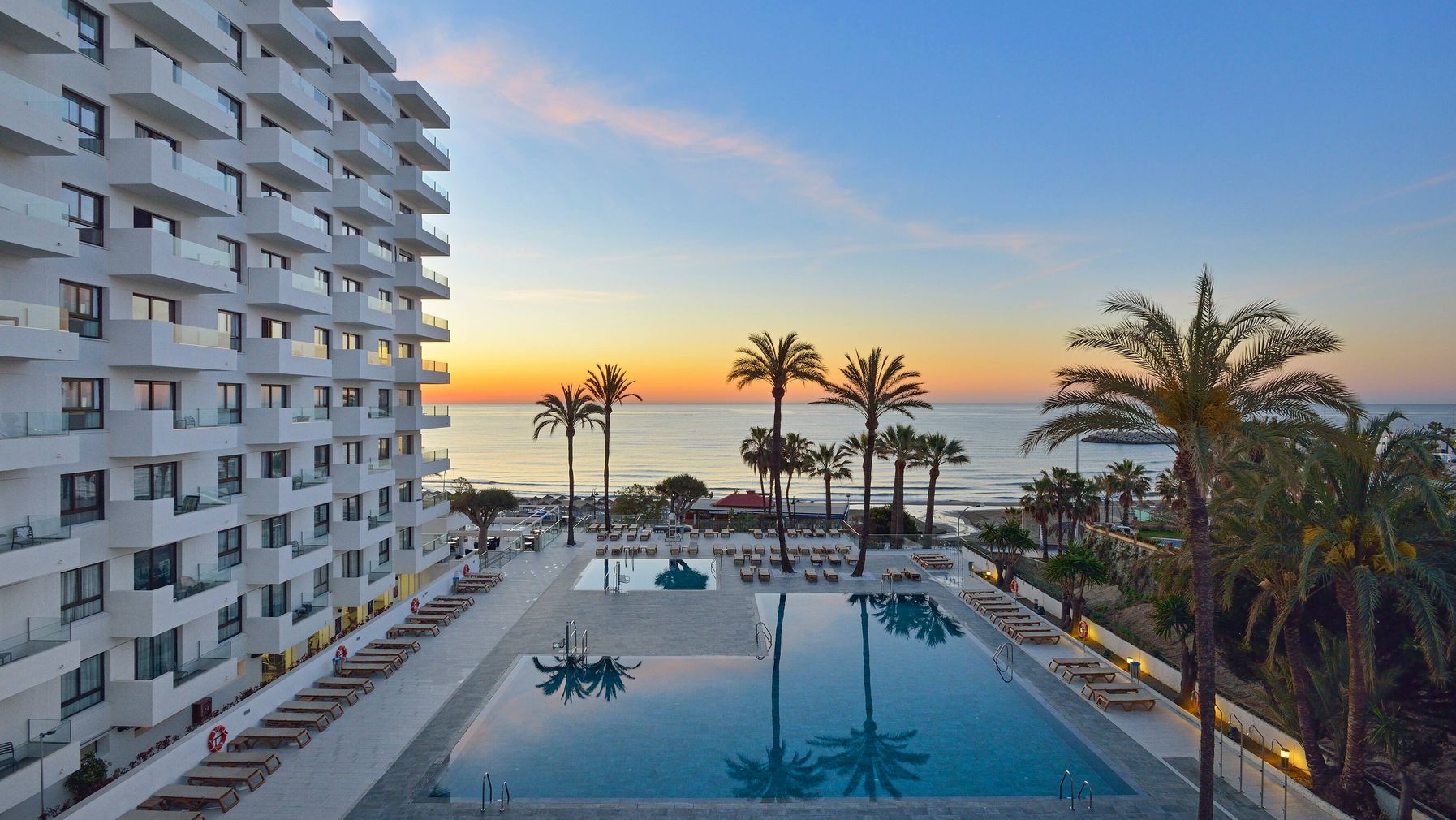 Ocean House Costa del Sol viesnīcas ēka un āra baseini saulrietā Torremolinosā, Spānijā — skats uz jūru un palmām