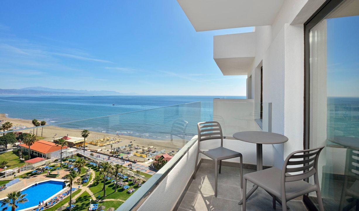 Skats no Ocean House Costa del Sol balkona uz smilšu pludmali, zilo jūru un viesnīcas dārzu ar baseinu