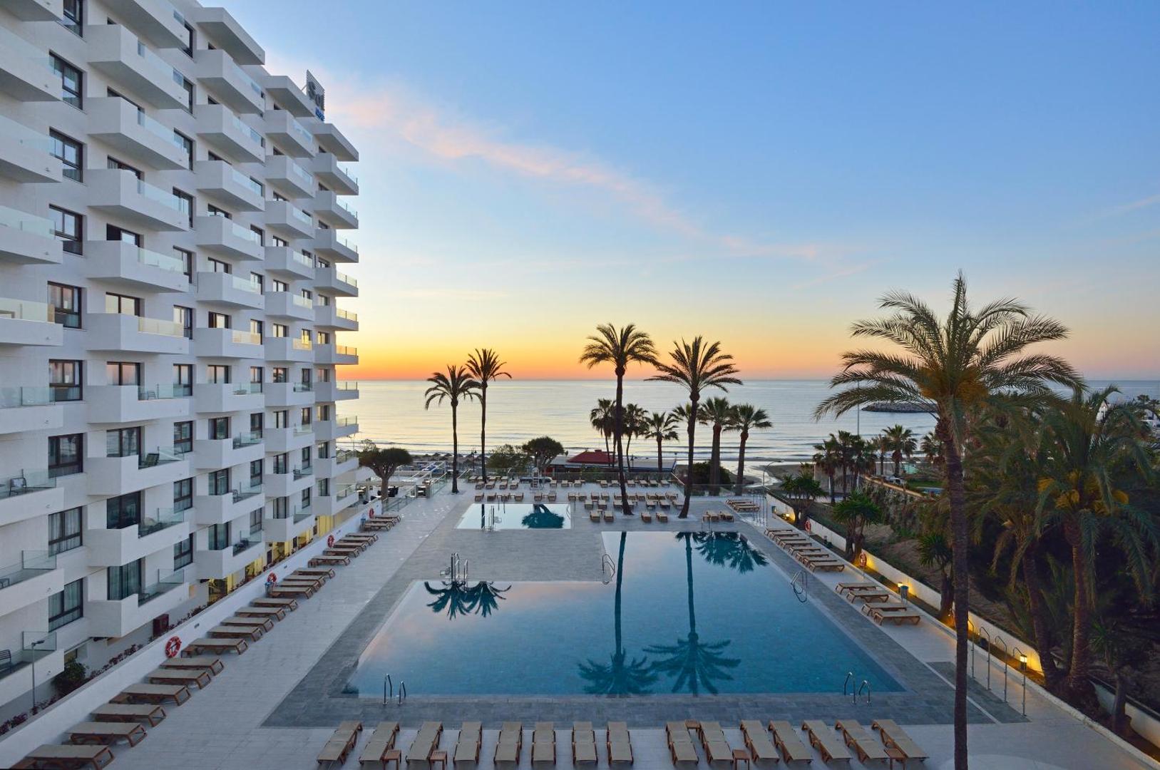 Ocean House Costa del Sol āra baseins saulrieta laikā ar skatu uz jūru un palmām — sauļošanās krēsli pie baseina malas