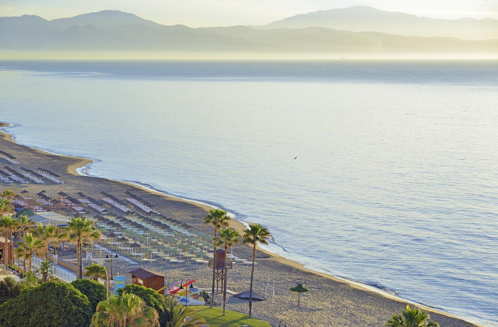 Plašs skats no Ocean House Costa del Sol uz smilšu pludmali, saulessargiem un kalniem tālumā rīta gaismā