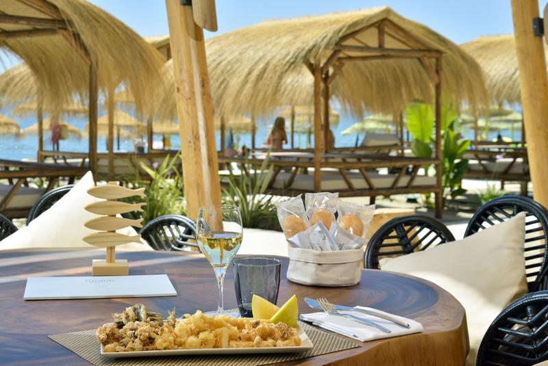 Melia Costa del Sol pludmales klubs ar salmu saulessargiem un gultām — galds ar jūras veltēm un vīnu priekšplānā