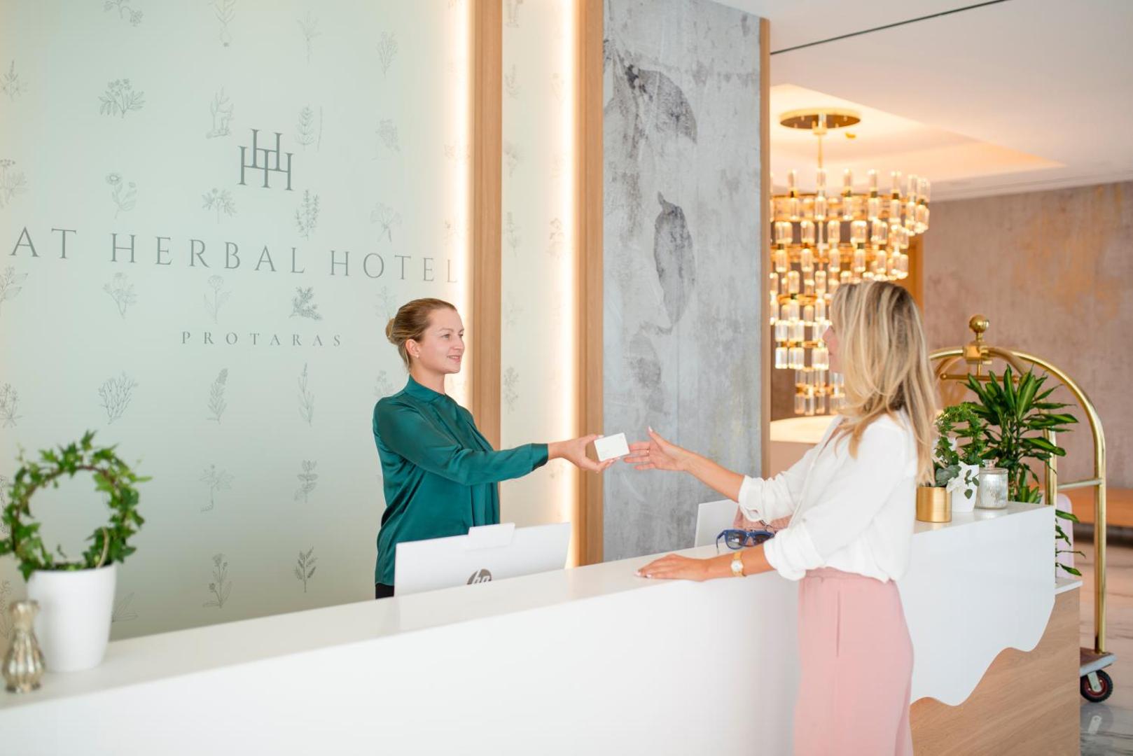 At Herbal Boutique Hotel & Spa reģistratūra Protarasā — viesmīlīga darbiniece pie baltas letes sniedz numura atslēgu viesim