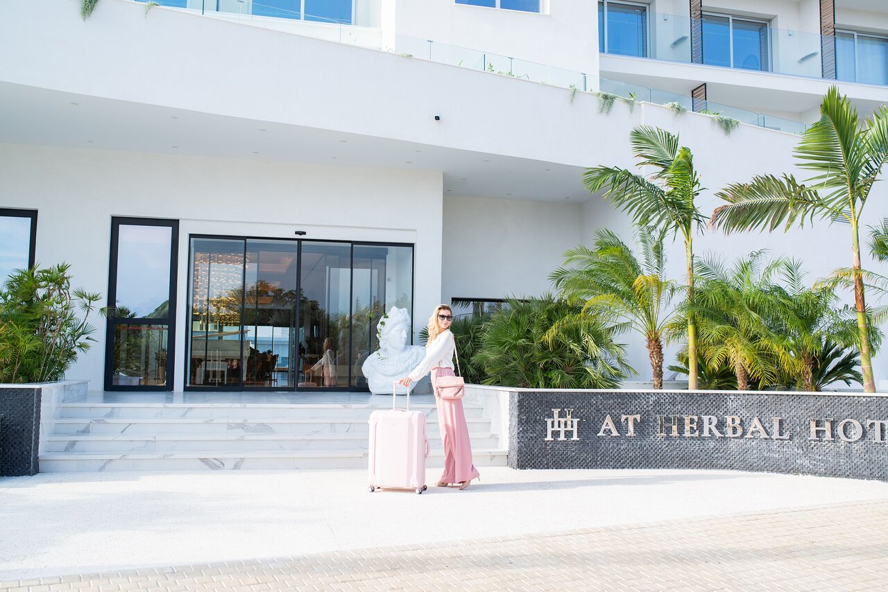 Viesis ar rozā koferi pie At Herbal Boutique Hotel & Spa ieejas Protarasā — gaiša, moderna arhitektūra un palmas