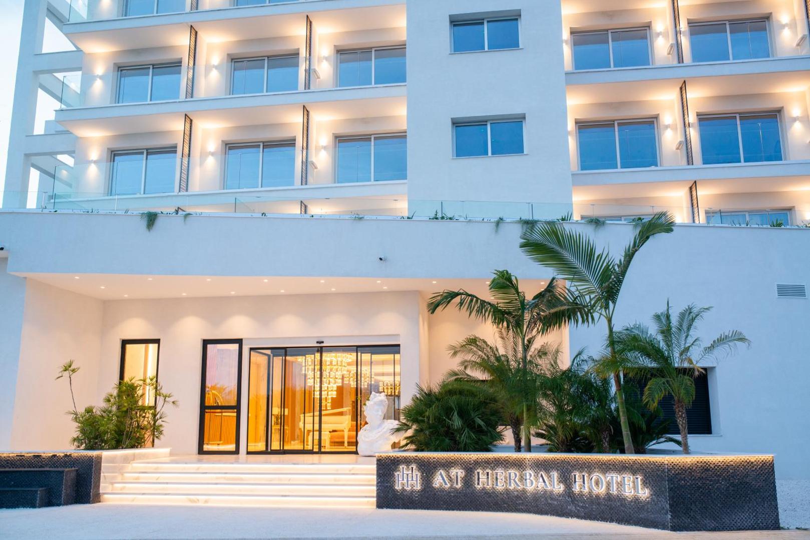 At Herbal Boutique Hotel & Spa galvenā ieeja Protarasā, Kiprā — moderna balta fasāde ar palmām un izgaismotu viesnīcas nosaukumu vakarā