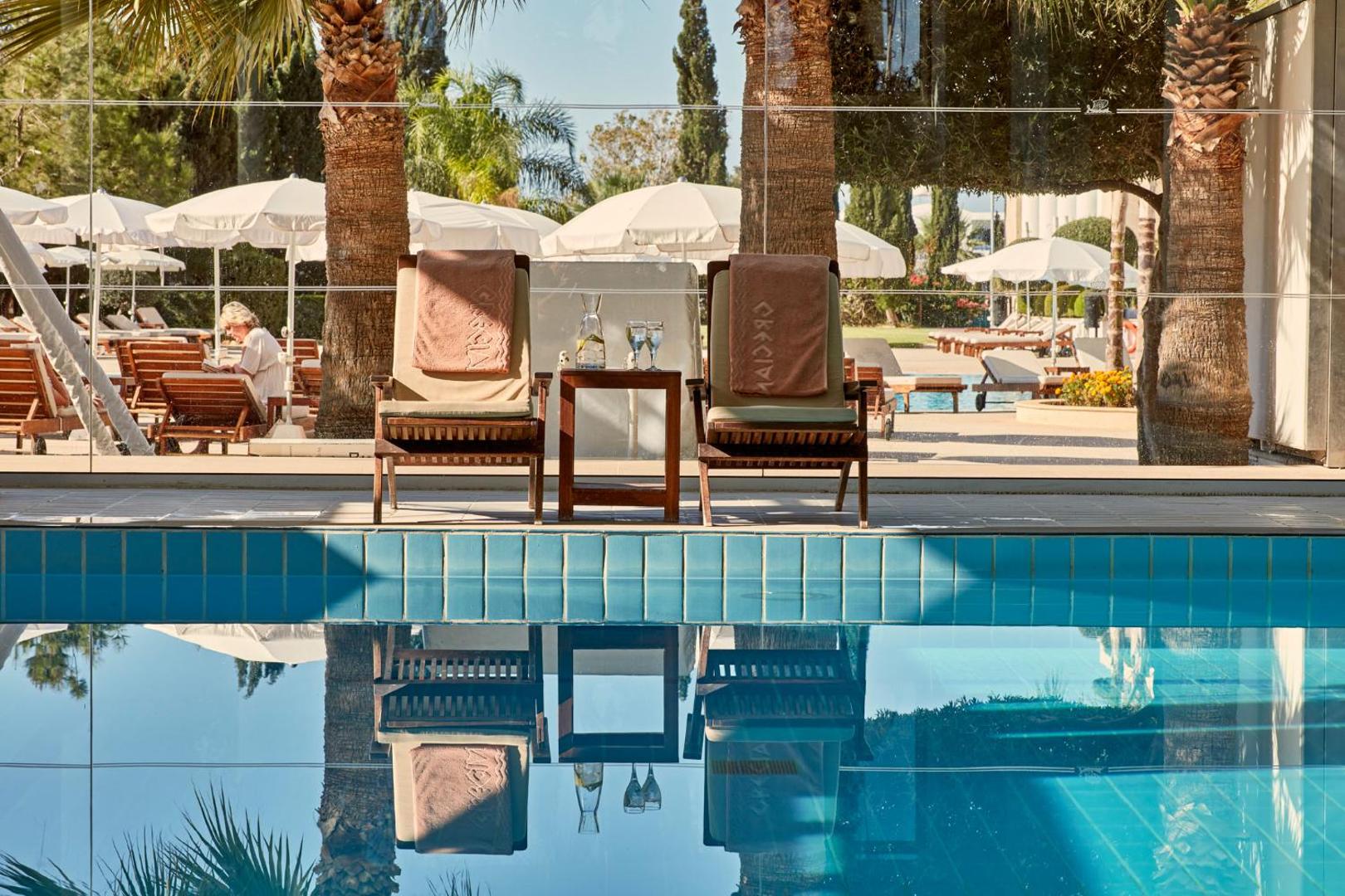Grecian Park baseina malas atpūtas zona ar koka krēsliem, dvieļiem un atspulgu ūdenī — fonā saulessargi un palmas Protarasā