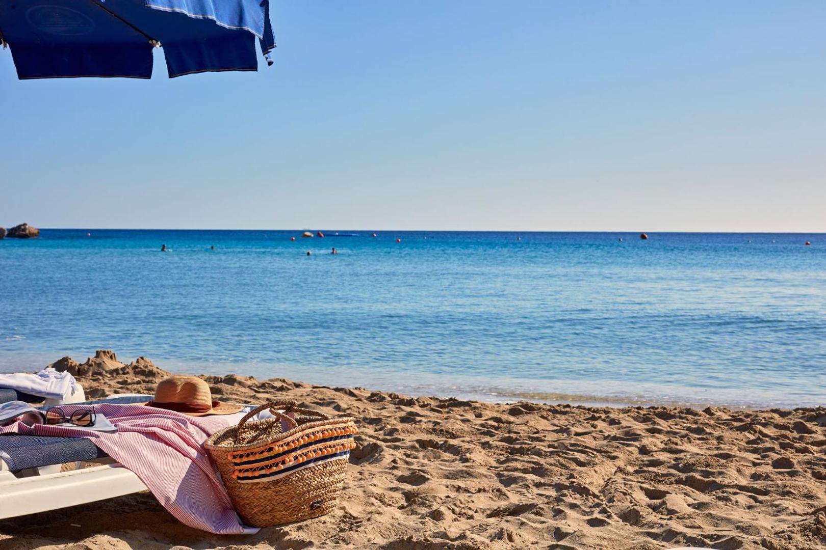 Grecian Park pludmales skats ar zilu saulessargu, pītu somu un cepuri uz sauļošanās krēsla pie dzidri zilas jūras Kiprā