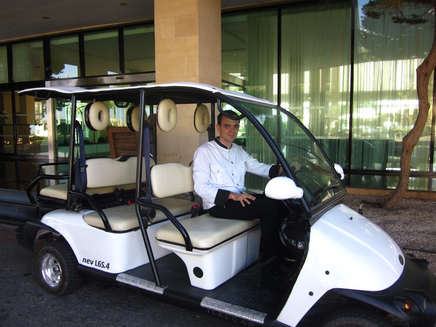 Grecian Park viesnīcas darbinieks pie balta golfa tipa elektroauto, kas paredzēts viesu pārvadāšanai teritorijā