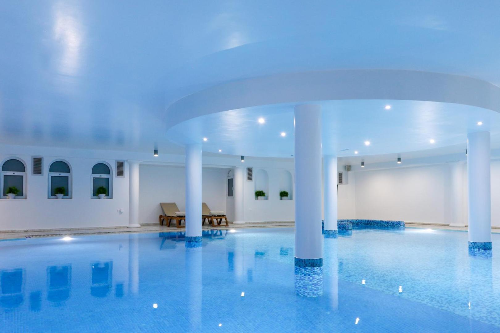 Golden Bay Beach iekštelpu baseins ar gaiši zilu apgaismojumu un kolonnām — relaksācijas zona ar atpūtas krēsliem viesnīcas spa centrā