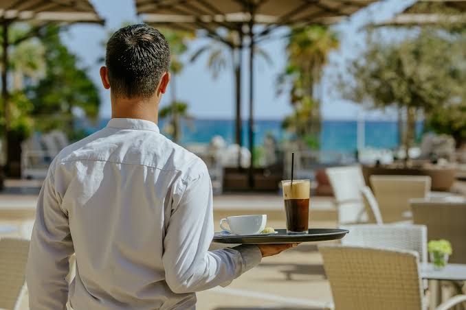 Viesmīlis ar dzērieniem uz paplātes Golden Bay Beach āra terasē ar skatu uz Vidusjūru