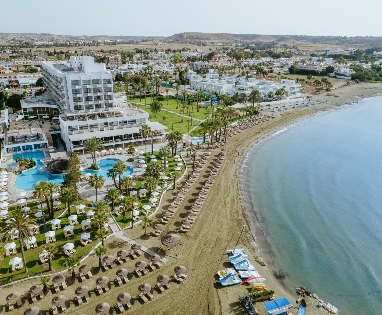 Golden Bay Beach viesnīcas ēka un plašā teritorija pie Vidusjūras krasta ar palmām, saulessargiem un ūdens sporta inventāru pludmalē.