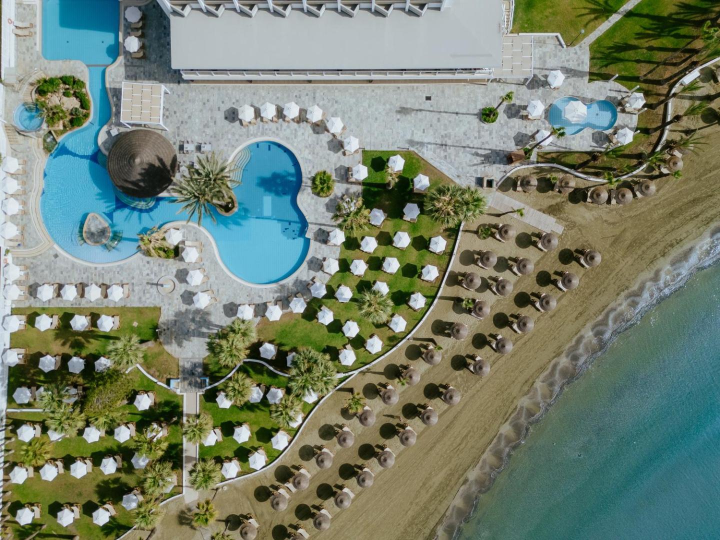 Golden Bay Beach viesnīcas skats no putna lidojuma Larnakā, Kiprā — āra baseini, sauļošanās krēsli ar saulessargiem un pieguļošā smilšu pludmale