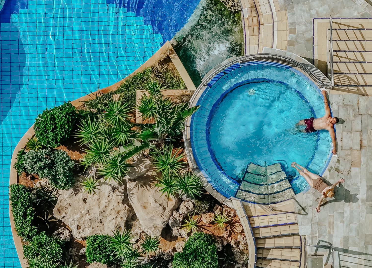 Skats no augšas uz Golden Bay Beach džakuzi un baseina zonu — viesi atpūšas ūdenī starp tropiskiem augiem un akmens dārzu