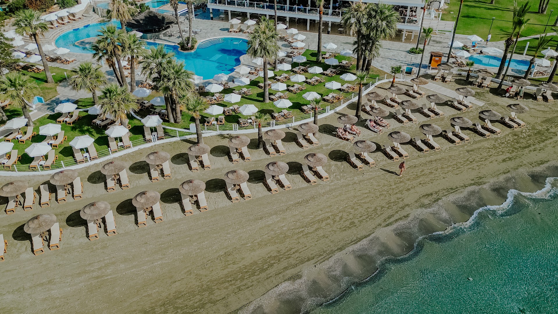 Aerofoto skats uz Golden Bay Beach teritoriju ar lielu āra baseinu, koptu zālienu un saulessargiem smilšu pludmalē.