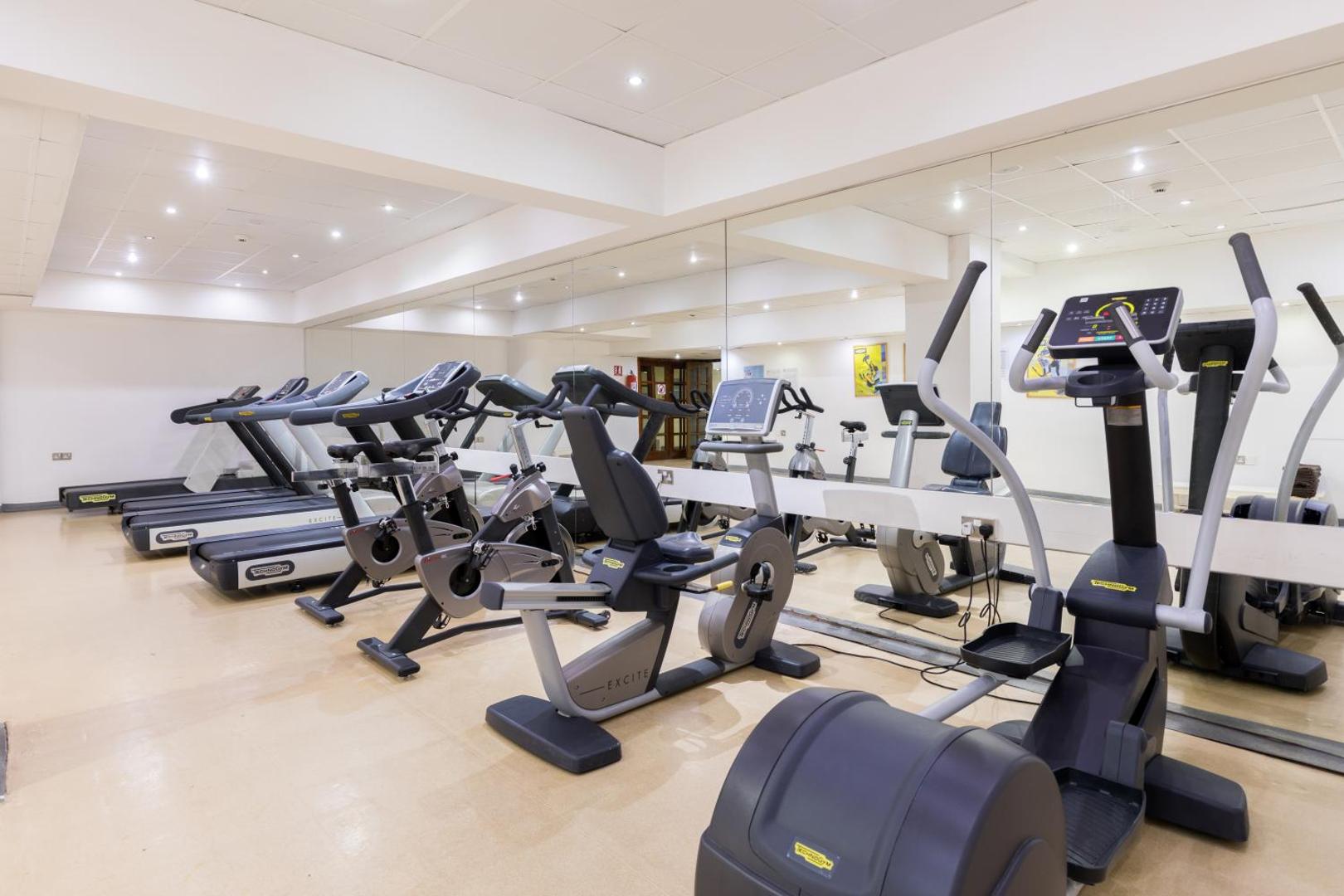 Golden Bay Beach viesnīcas fitnesa centrs Larnakā ar moderniem Technogym skrejceliņiem un velotrenažieriem gaišā telpā ar spoguļu sienu