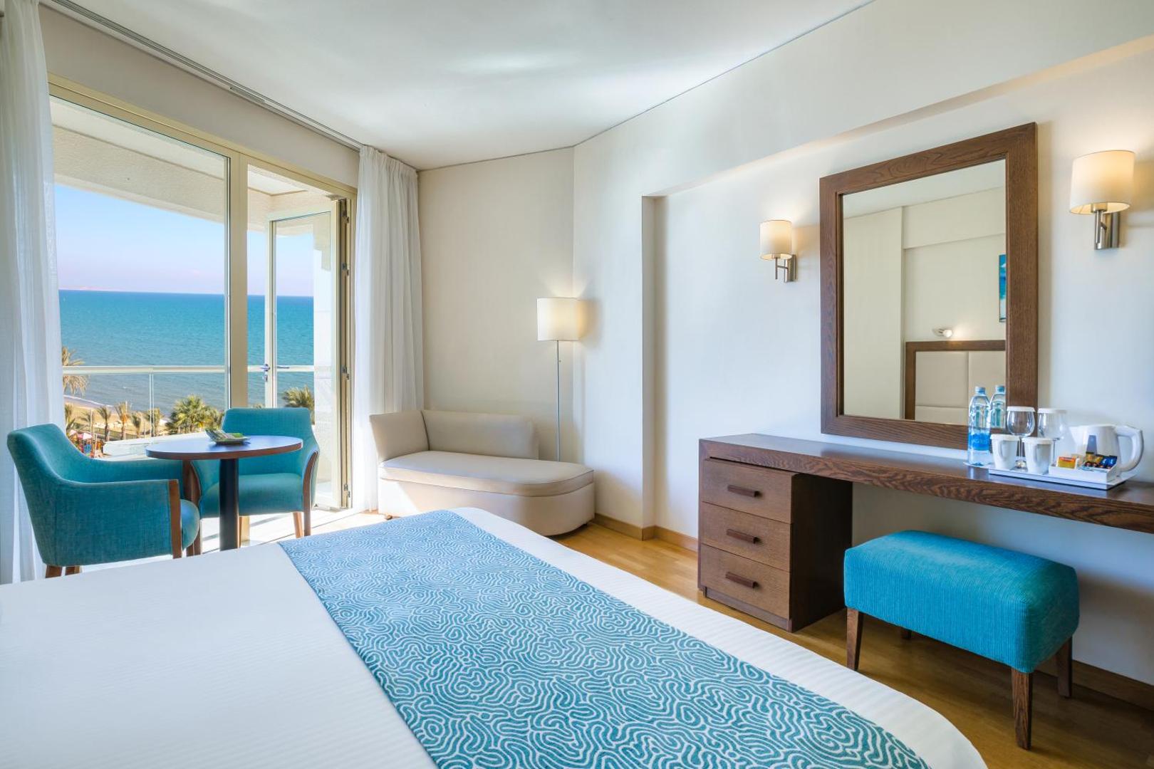Gaišs Golden Bay Beach viesnīcas numurs ar skatu uz jūru, ziliem akcentiem mēbelēs un balkonu Larnakā.
