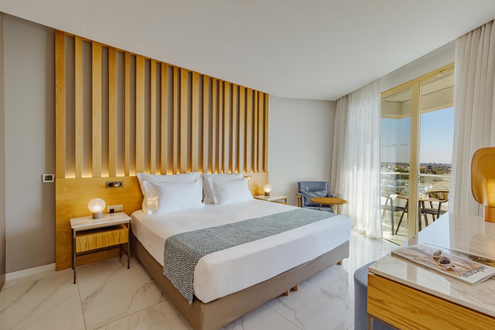 Moderns Golden Bay Beach viesnīcas numurs ar koka apdari, lielu gultu un gaišu interjeru Larnakas kūrortā.