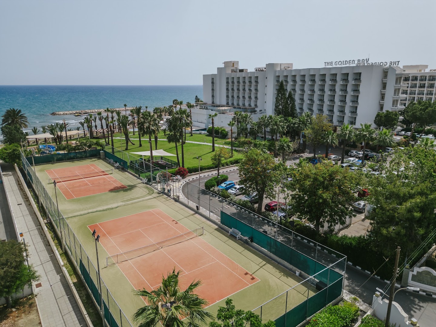 Golden Bay Beach viesnīcas ēka un divi tenisa korti Larnakas piekrastē — skats no augšas uz dārzu, palmām un Vidusjūru