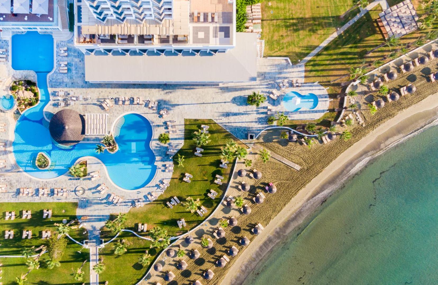 Golden Bay Beach viesnīcas skats no putna lidojuma uz āra baseiniem, sauļošanās zonu ar zālienu un smilšu pludmali Larnakā, Kiprā.