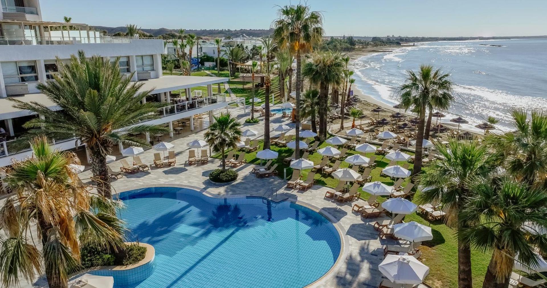 Golden Bay Beach viesnīcas teritorija Larnakā — liels āra baseins, palmas un sauļošanās krēsli tieši pie smilšu pludmales līča