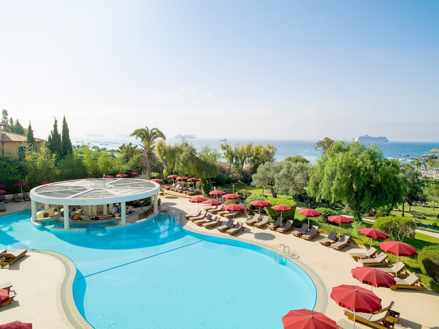 St. Raphael Resort āra baseins ar bāru ūdenī un skatu uz Vidusjūru Limasolā, Kiprā — atpūtas zona ar sarkaniem saulessargiem