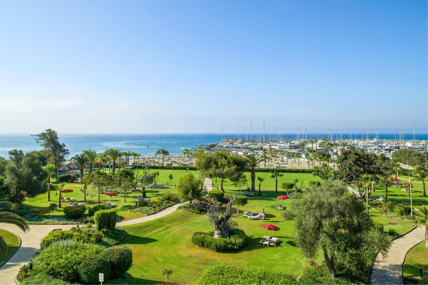 St. Raphael Resort plašā dārza panorāma Limasolā ar skatu uz jahtu piestātni, Vidusjūru un zaļo atpūtas zonu ar palmām