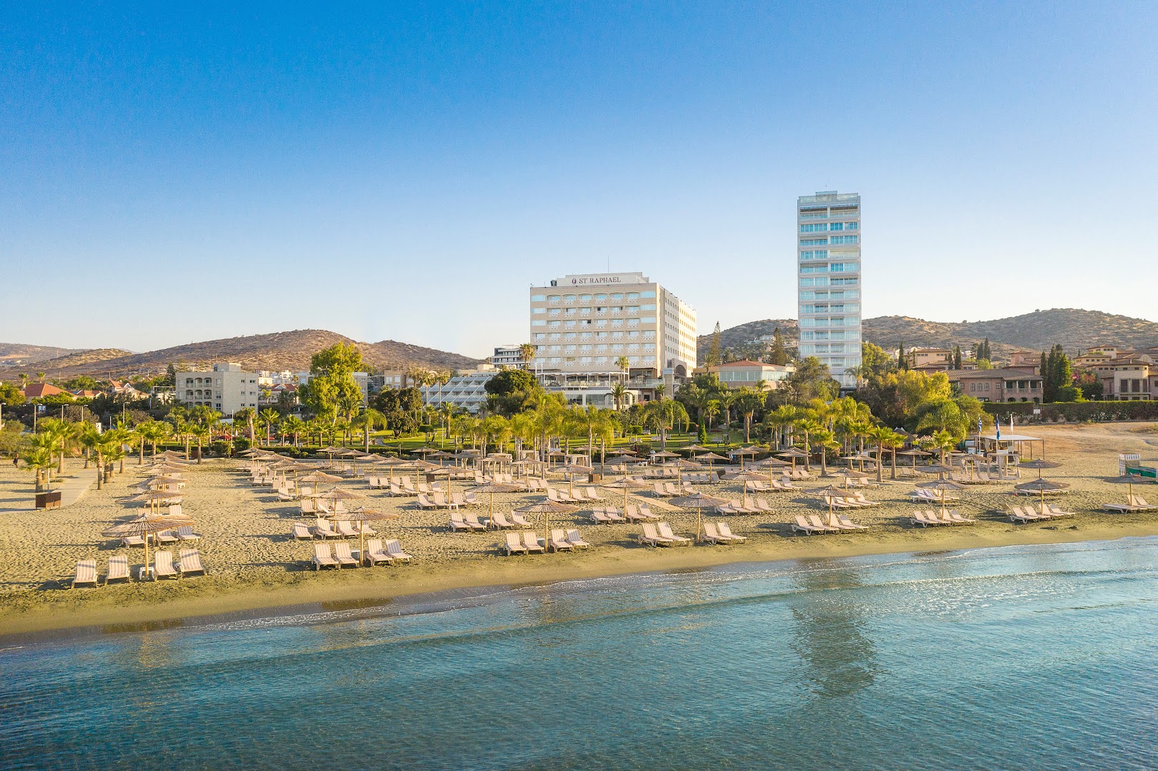 Viesnīcas St. Raphael Resort galvenā ēka un pludmales zona Limasolā — skats no jūras puses uz kūrorta fasādi un saulessargiem