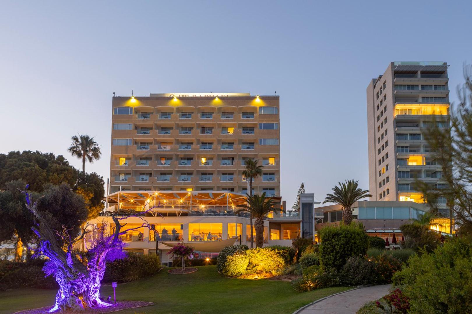 St. Raphael Resort galvenā ēka un dārzs vakara apgaismojumā Limasolā — izgaismotas terases un labiekārtota teritorija krēslas stundā