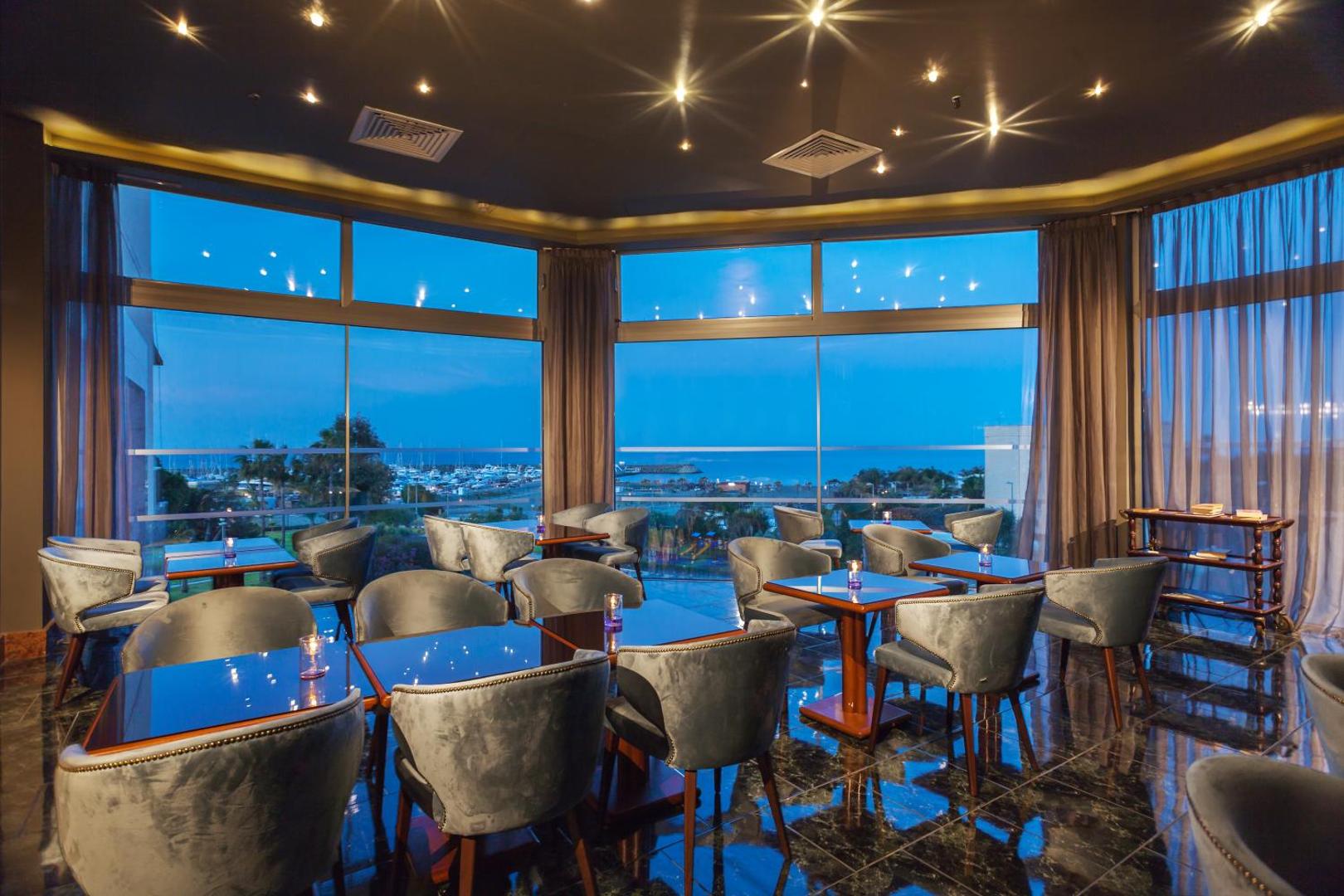 St. Raphael Resort restorāns ar panorāmas logiem uz Limasolas jahtu piestātni vakarā, ziliem samta krēsliem un sveču gaismu