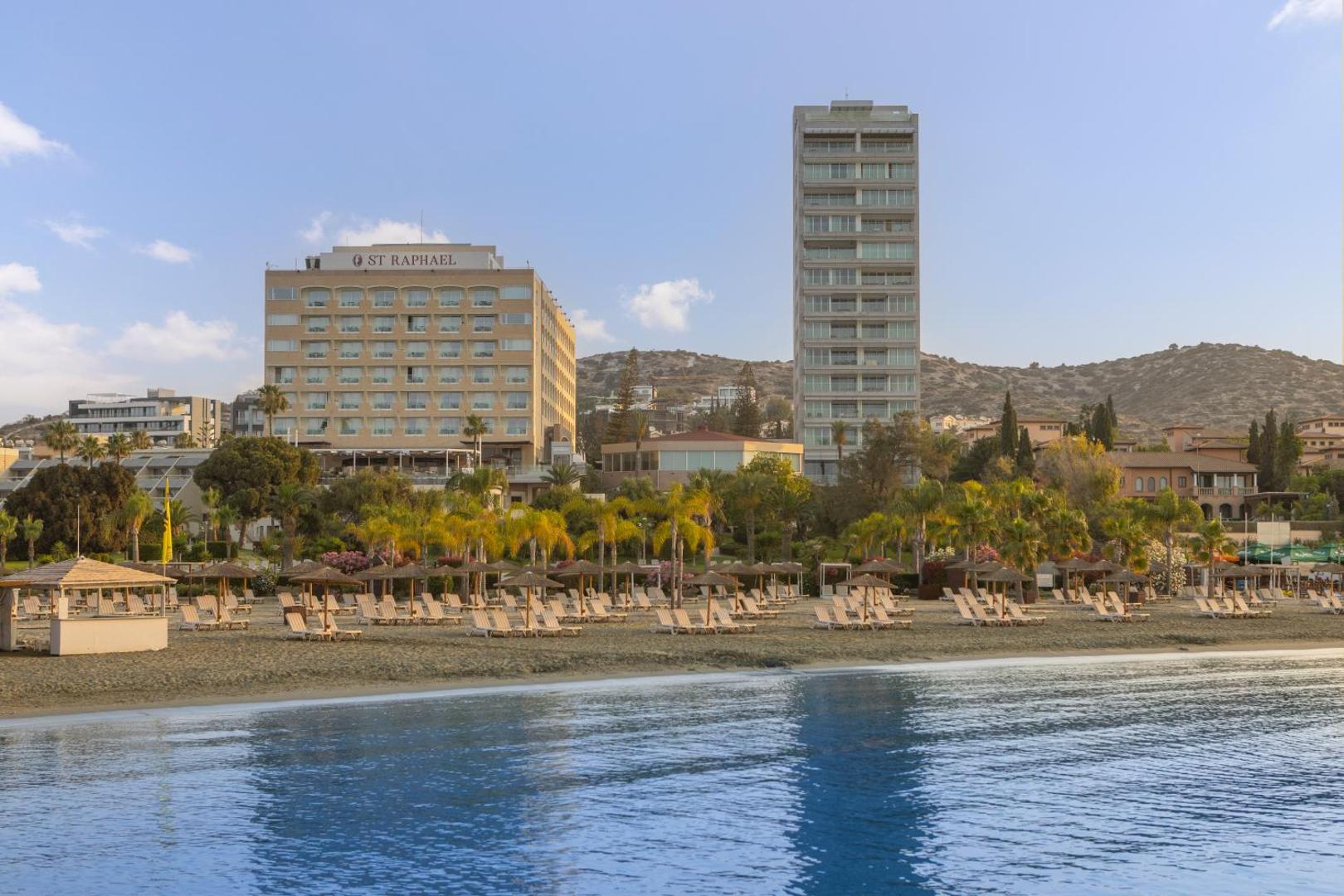 St. Raphael Resort skats no jūras puses Limasolā, Kiprā — smilšu pludmale ar saulessargiem un viesnīcas ēkas palmu ieskāvumā