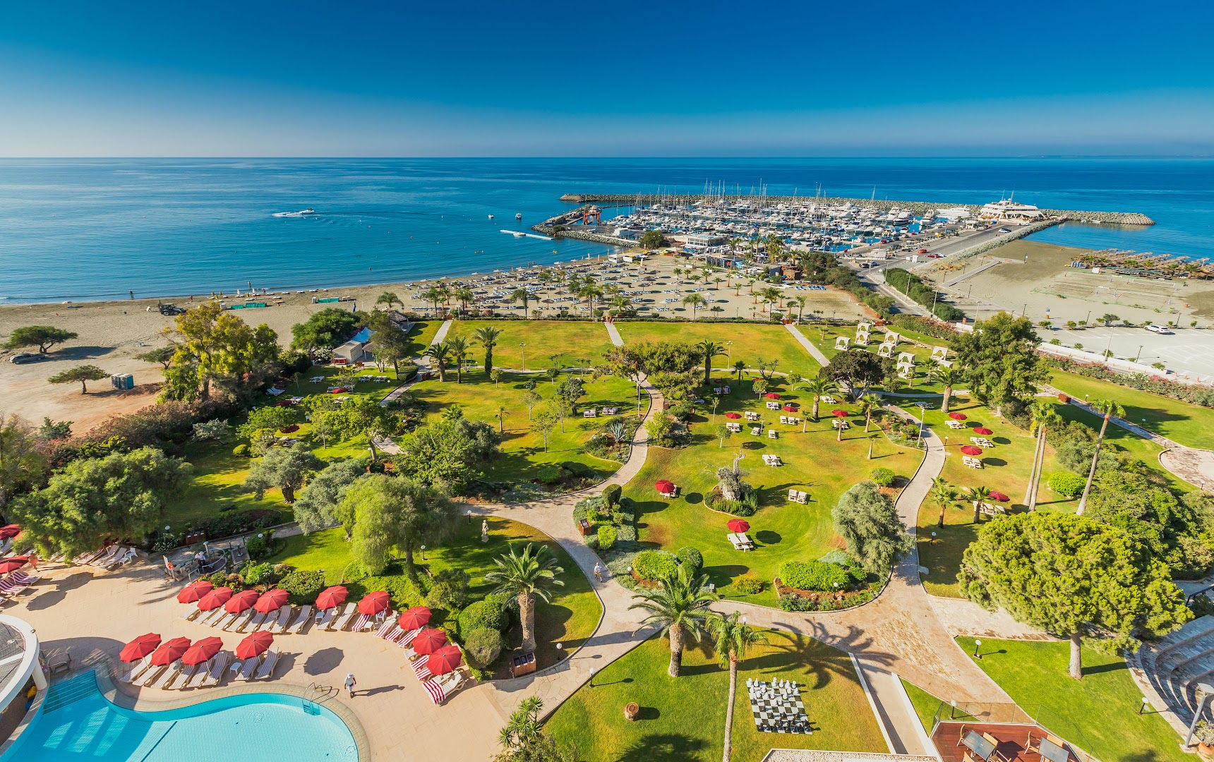 St. Raphael Resort plašā dārza teritorija ar gājēju celiņiem, palmām un skatu uz pludmali un jahtu piestātni Limasolā