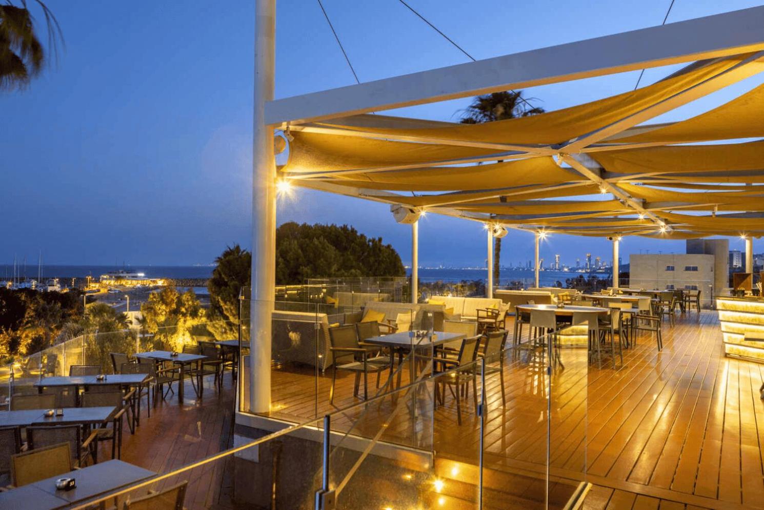 St. Raphael Resort āra terase vakarā ar modernu apgaismojumu, koka klāju un skatu uz naksnīgo Limasolas piekrasti