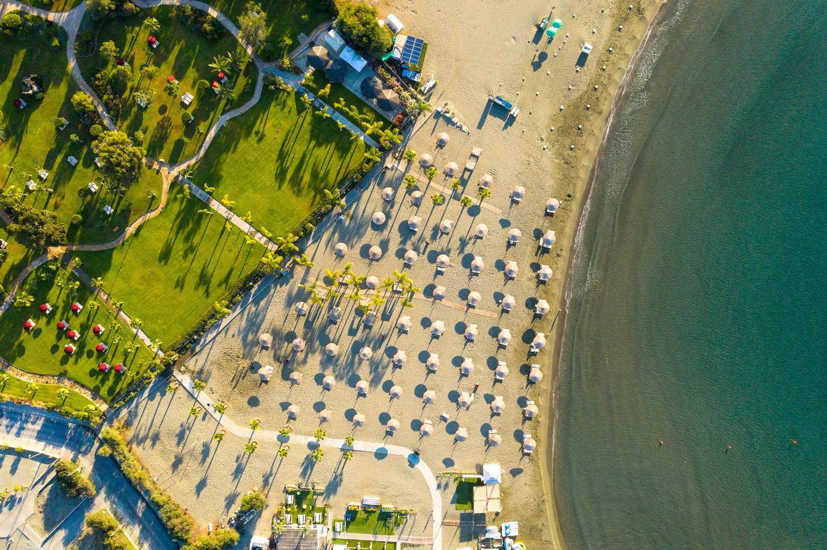 St. Raphael Resort skats no putna lidojuma uz pludmali un zaļo dārzu Limasolā, Kiprā — saulessargi, palmas un jūras krasts