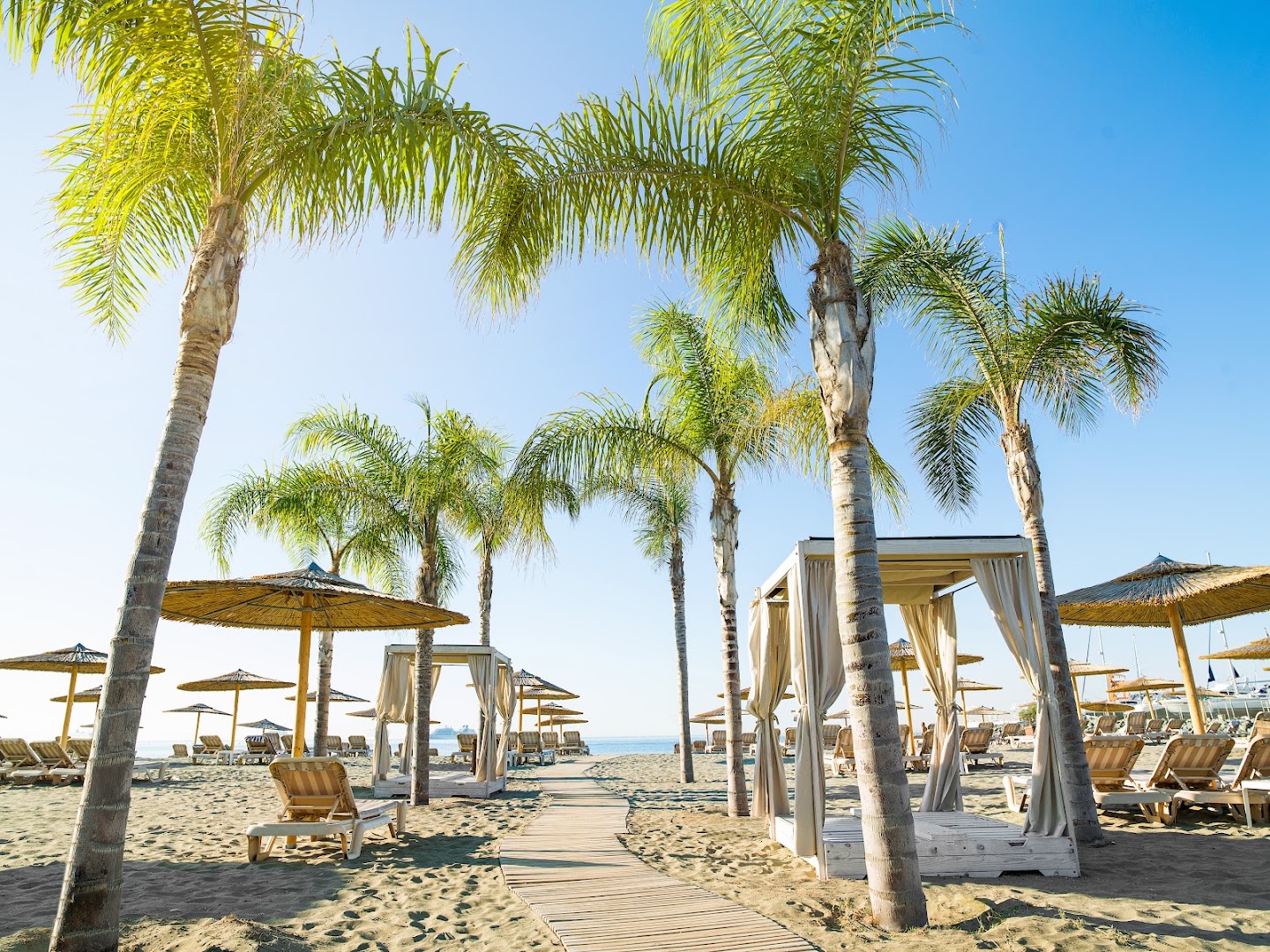 St. Raphael Resort pludmales zona ar palmām, koka laipām un atpūtas nojumēm — gaišas smiltis un saulains laiks Limasolā