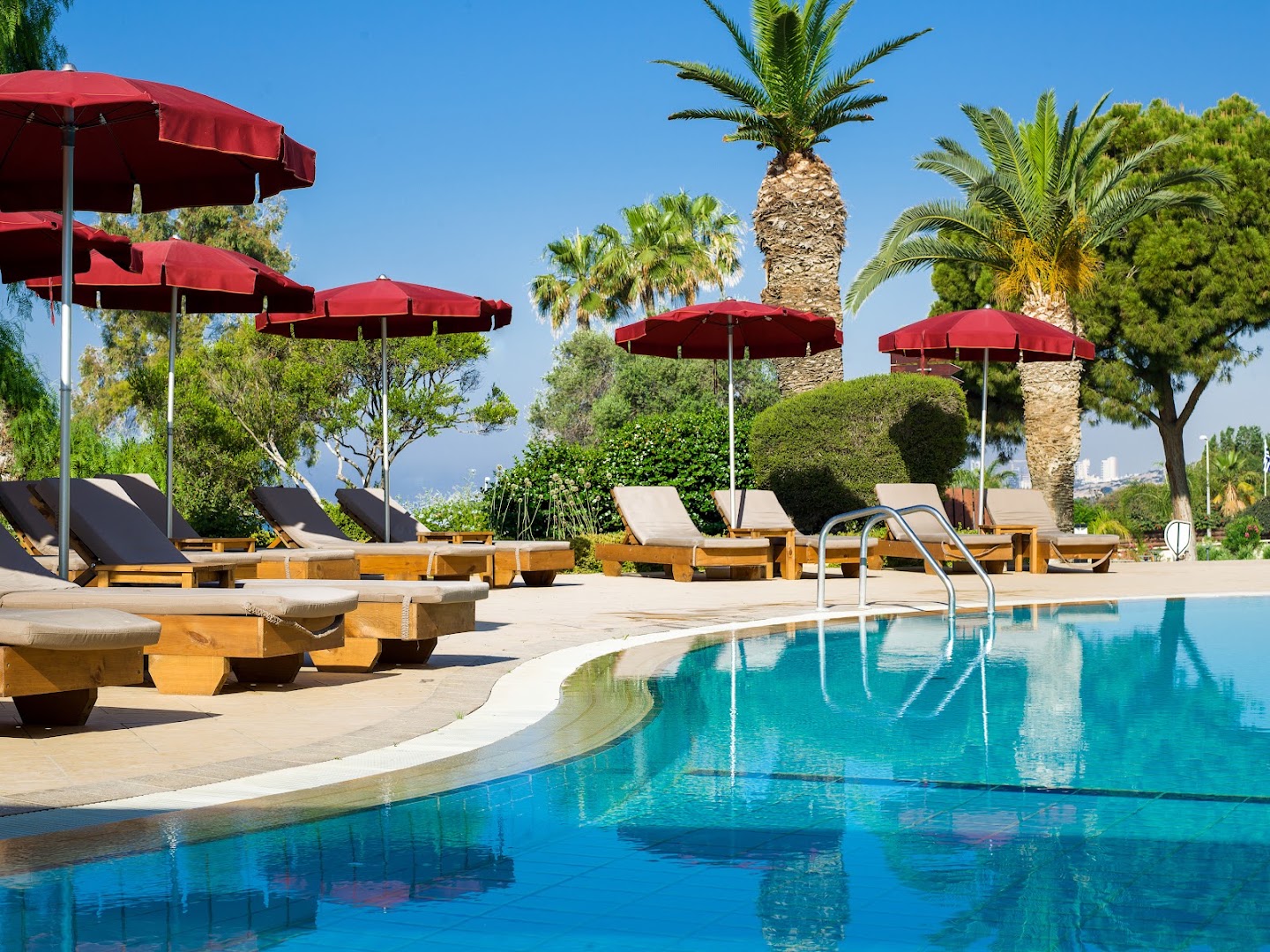 St. Raphael Resort sauļošanās krēsli un sarkani saulessargi pie dzidra baseina Limasolā — fonā palmas un zilas debesis