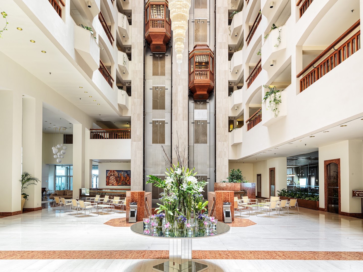 St. Raphael Resort plašais ātrija vestibils Limasolā ar stikla liftiem, koka apdari, ziedu kompozīcijām un atpūtas zonām vairākos stāvos