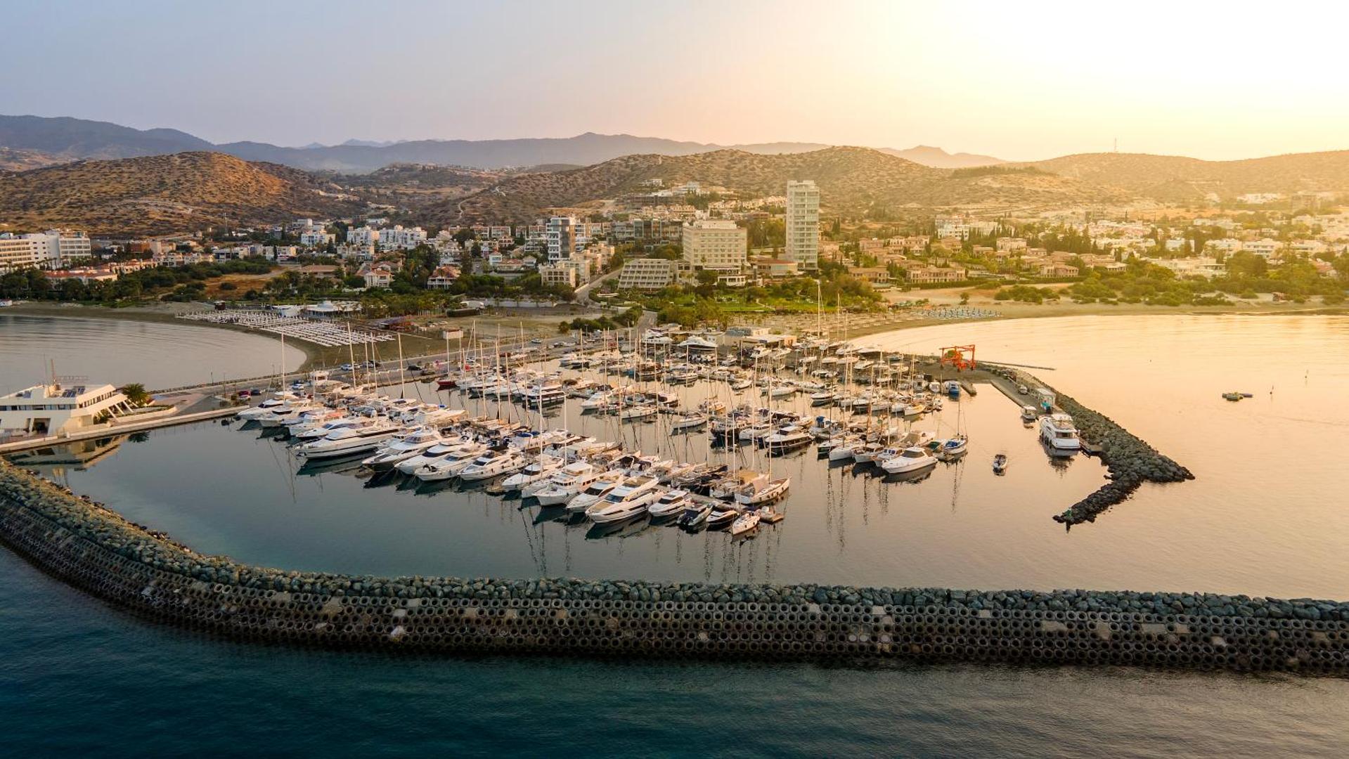 Aerofoto skats uz St. Raphael Resort jahtu piestātni un viesnīcas ēkām Limasolā saulrieta laikā — kalni fonā