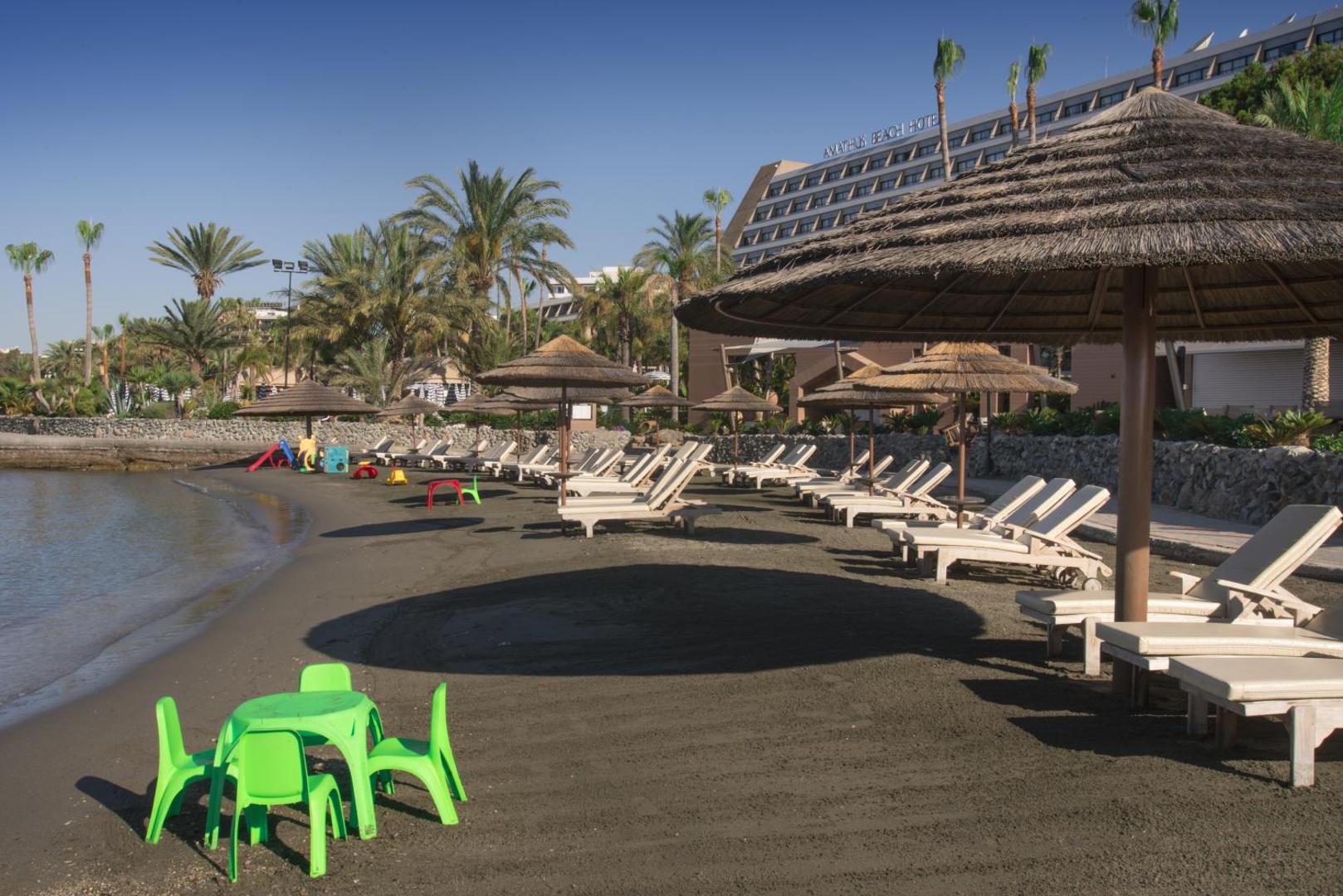 Amathus Beach Hotel smilšu pludmale ar sauļošanās krēsliem, salmu saulessargiem un bērnu mēbelēm uz viesnīcas ēkas fona