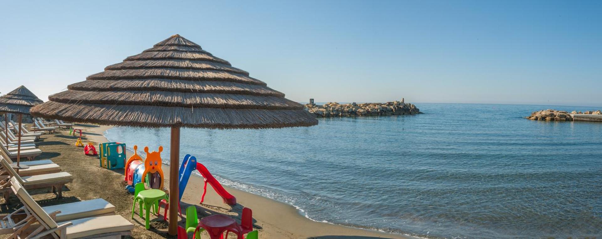 Amathus Beach Hotel bērnu rotaļu laukums pludmalē — krāsaini slidkalniņi un mēbeles zem salmu saulessarga pie jūras