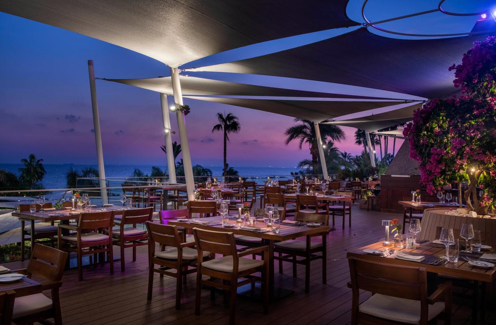 Amathus Beach Hotel āra restorāna terase vakarā — klāti galdi zem nojumēm ar skatu uz saulrietu un jūru