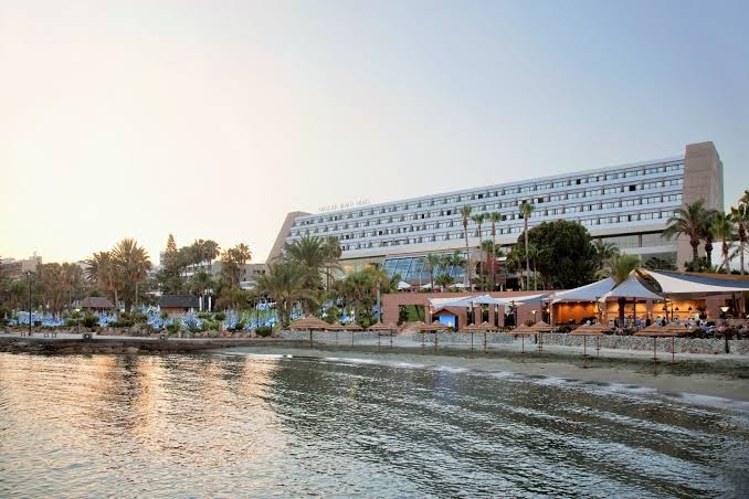 Amathus Beach Hotel skats no jūras puses uz viesnīcas ēku un pludmales bāru saulrietā Limasolā