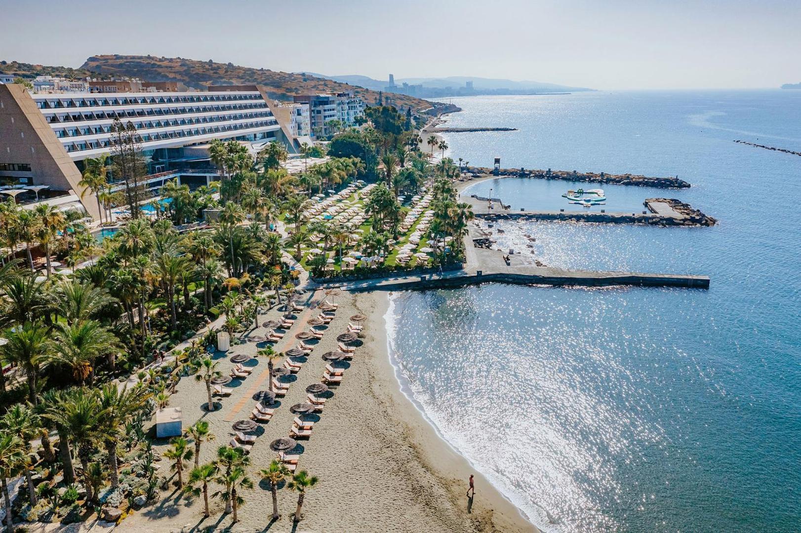 Amathus Beach Hotel panorāmas skats Limasolā, Kiprā — viesnīcas ēka, palmu dārzs un smilšu pludmale pie Vidusjūras līča