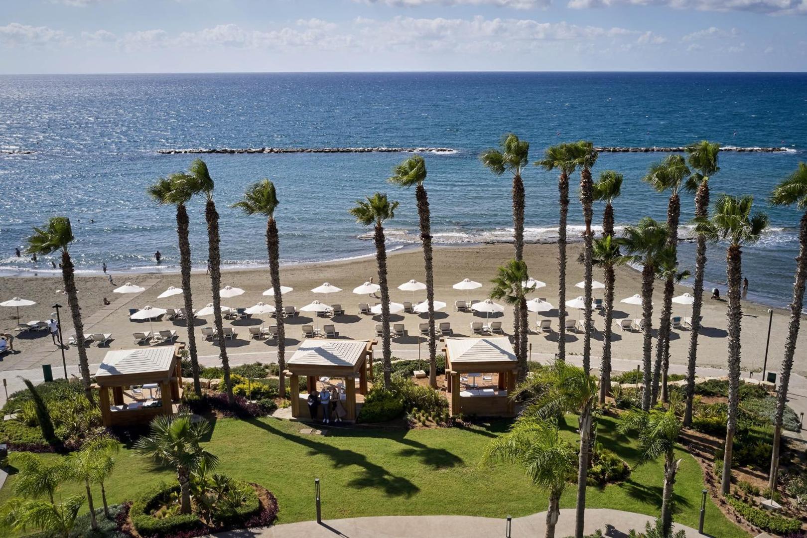 Parklane Resort & Spa skats no augšas uz pludmali Limasolā, Kiprā — smilšu krasts ar saulessargiem, palmām un atpūtas nojumēm pie jūras