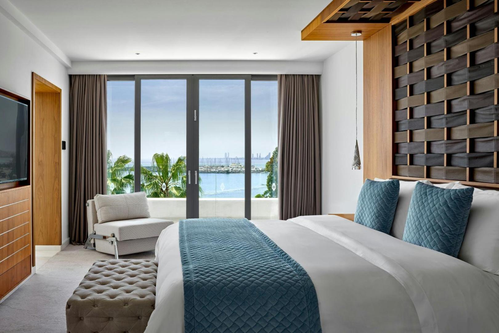 Parklane Resort & Spa numurs ar skatu uz jūru — gaišs interjers, liela gulta un panorāmas logs uz Limasolas jahtu piestātni